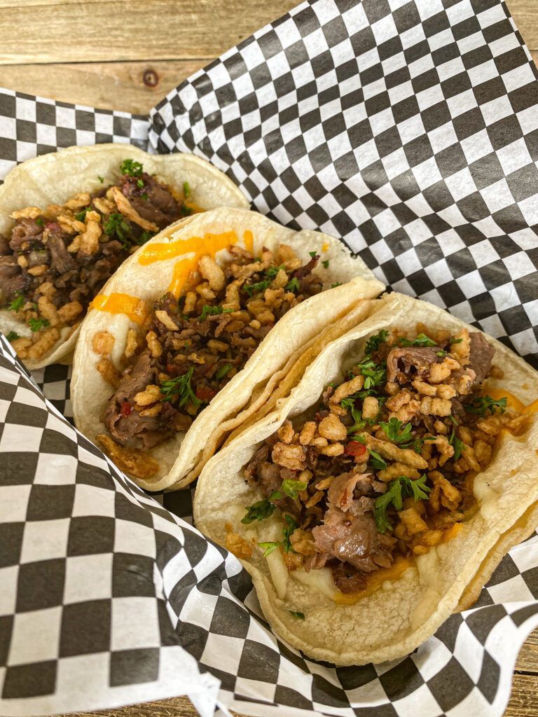 Three street prime rib tacos served on a black and white checked paper with a wood board in the background.